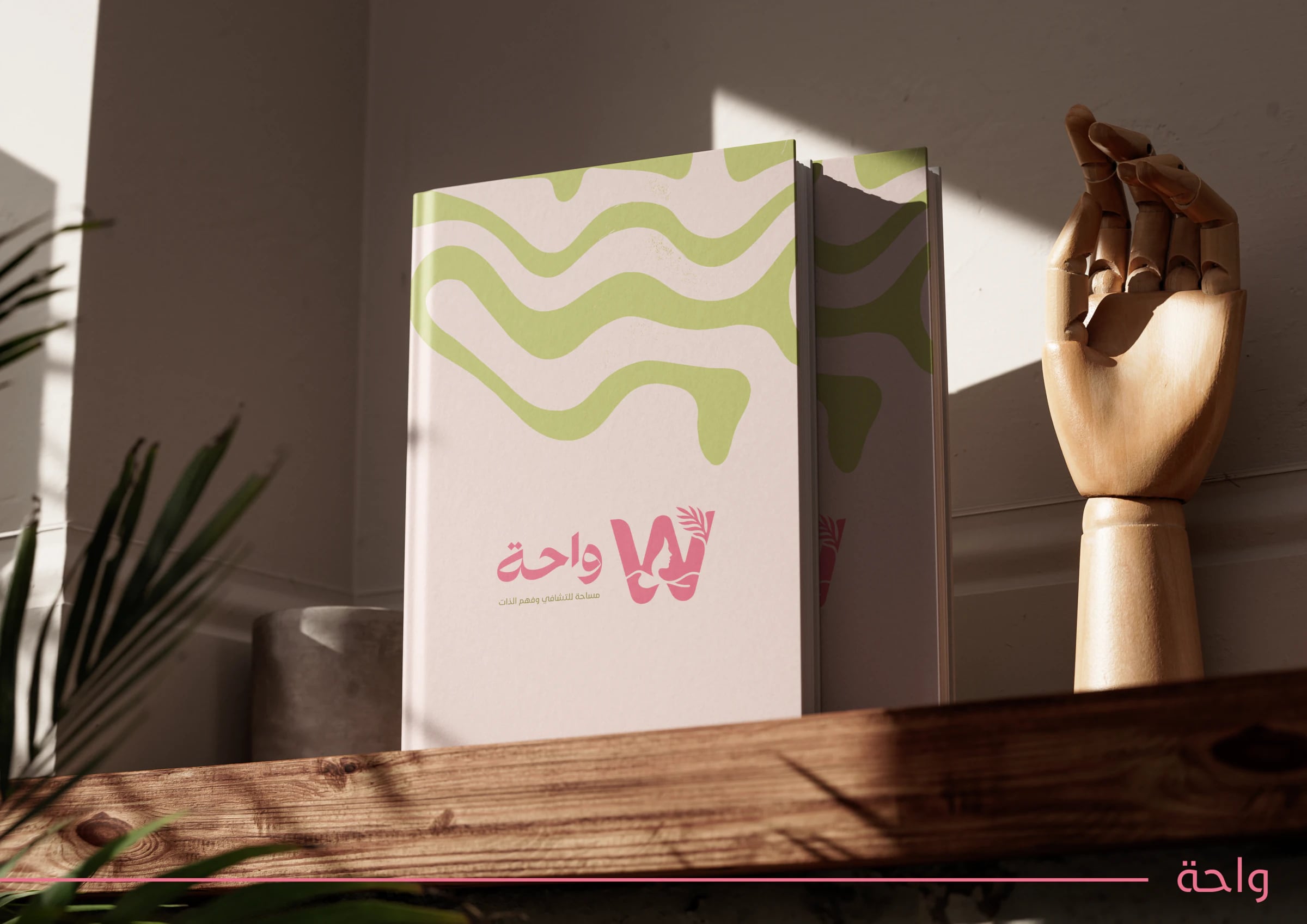 كتاب بغلاف وردي وأخضر منقوش عليه الشعار، بجانب مجسم يد خشبي في ضوء دافئ.